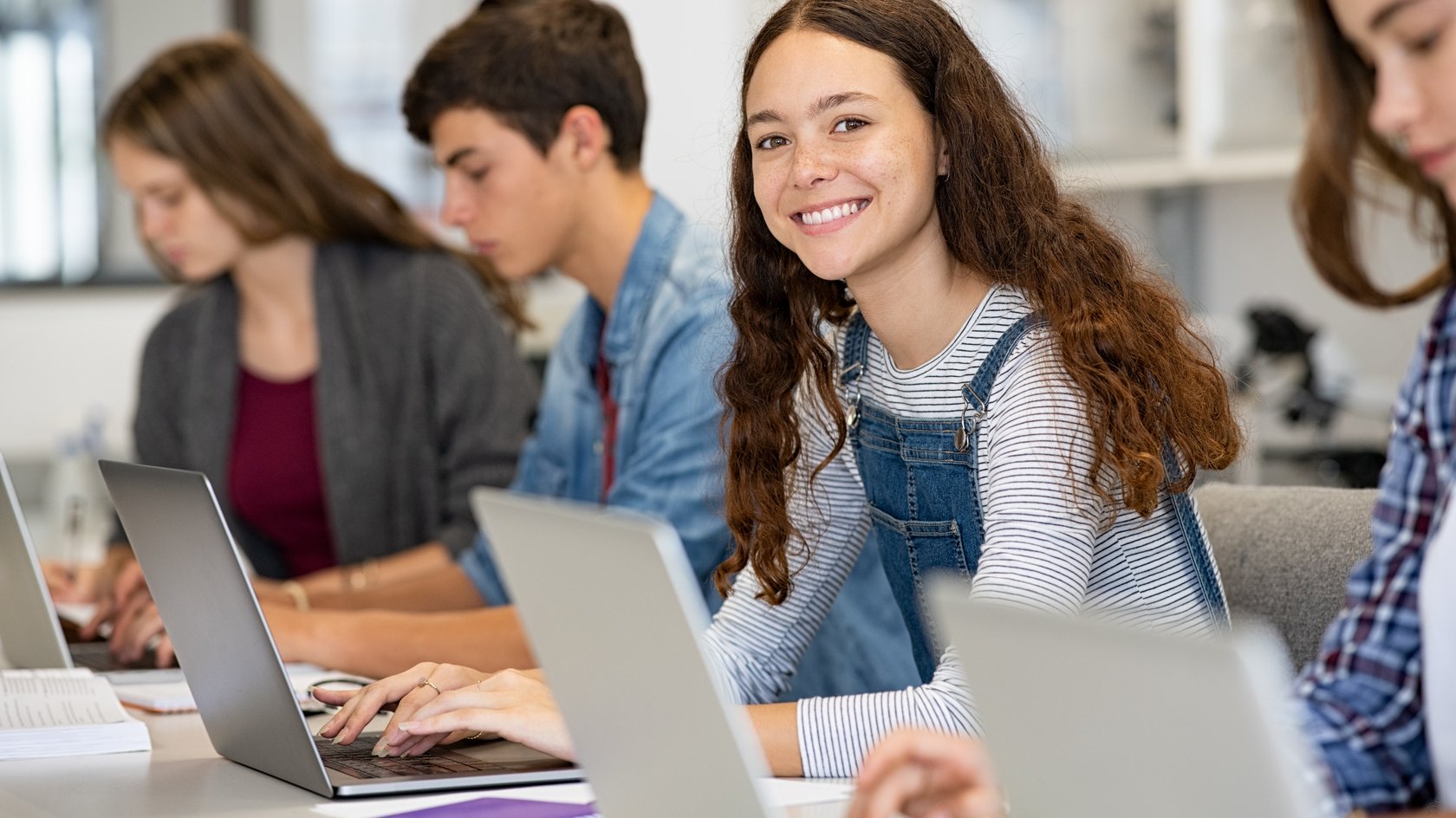 zwei Schülerinnen und zwei Schüler arbeiten konzentriert am Laptop. Eine Schülerin lächelt in die Kamera.