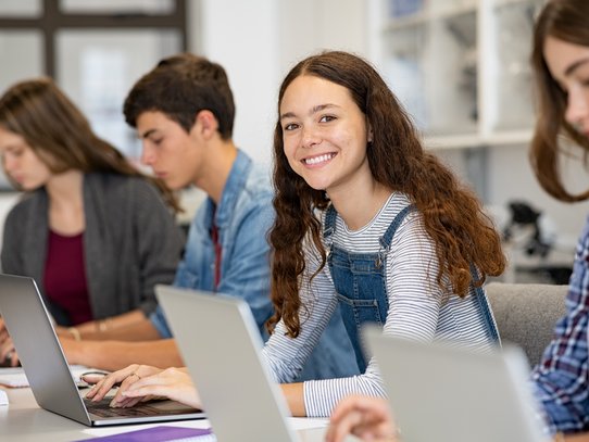 zwei Schülerinnen und zwei Schüler arbeiten konzentriert am Laptop. Eine Schülerin lächelt in die Kamera.