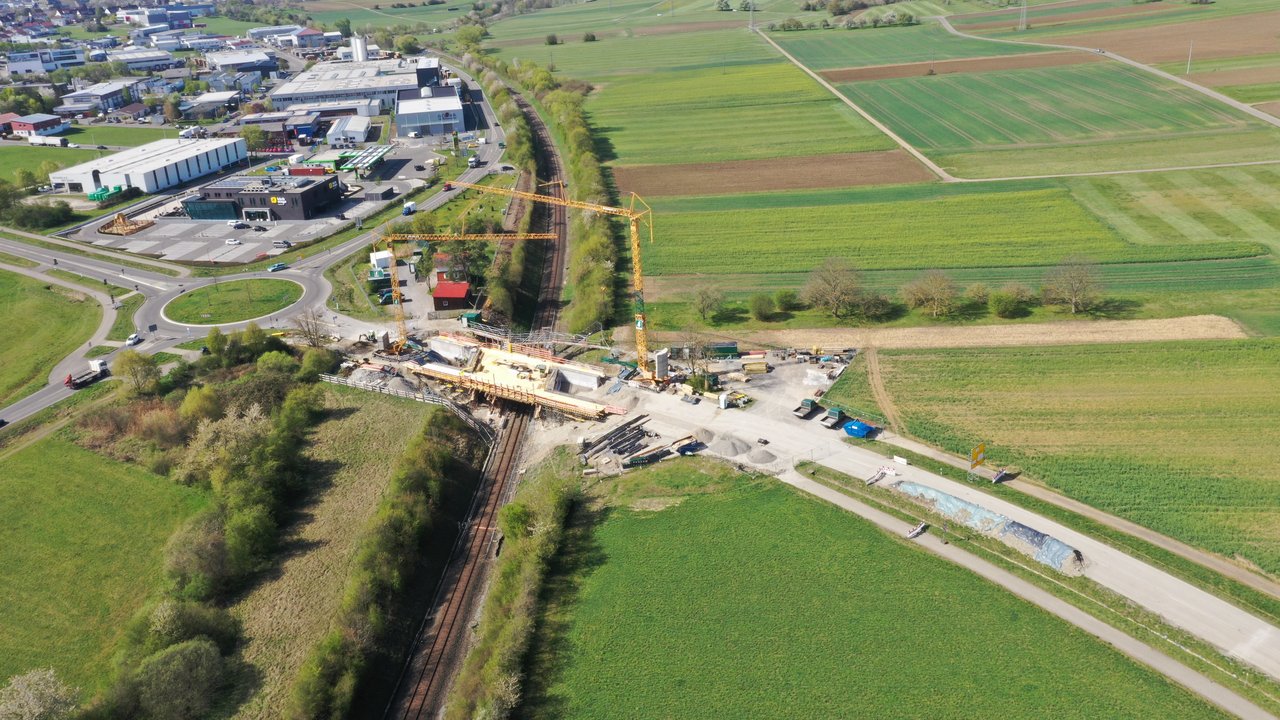 Luftbild einer Brückenbaustelle. In der Mitte des Bildes verläuft von vorne nach hinten eine Bahntrasse. Von links nach rechts läuft eine Straße. Am Schnittpunkt befindet sich eine Brücke, die die Straße über die Gleise führt. An dieser Brücke finden Arbeiten statt, zwei Kräne auf beiden Seiten heben Material.