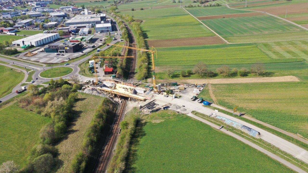 Luftbild einer Brückenbaustelle. In der Mitte des Bildes verläuft von vorne nach hinten eine Bahntrasse. Von links nach rechts läuft eine Straße. Am Schnittpunkt befindet sich eine Brücke, die die Straße über die Gleise führt. An dieser Brücke finden Arbeiten statt, zwei Kräne auf beiden Seiten heben Material.