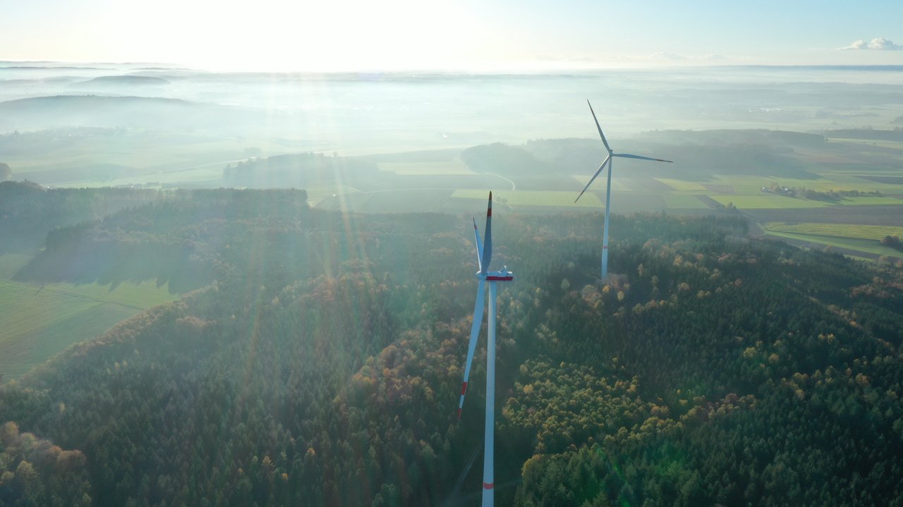 Das Foto zeigt zwei Windräder von oben inmitten einer grünen bewaldeten Landschaft