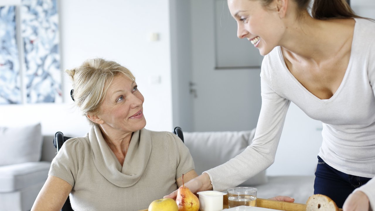 Junge Frau reicht einer Dame im Rollstuhl ein Tablett mit Essen