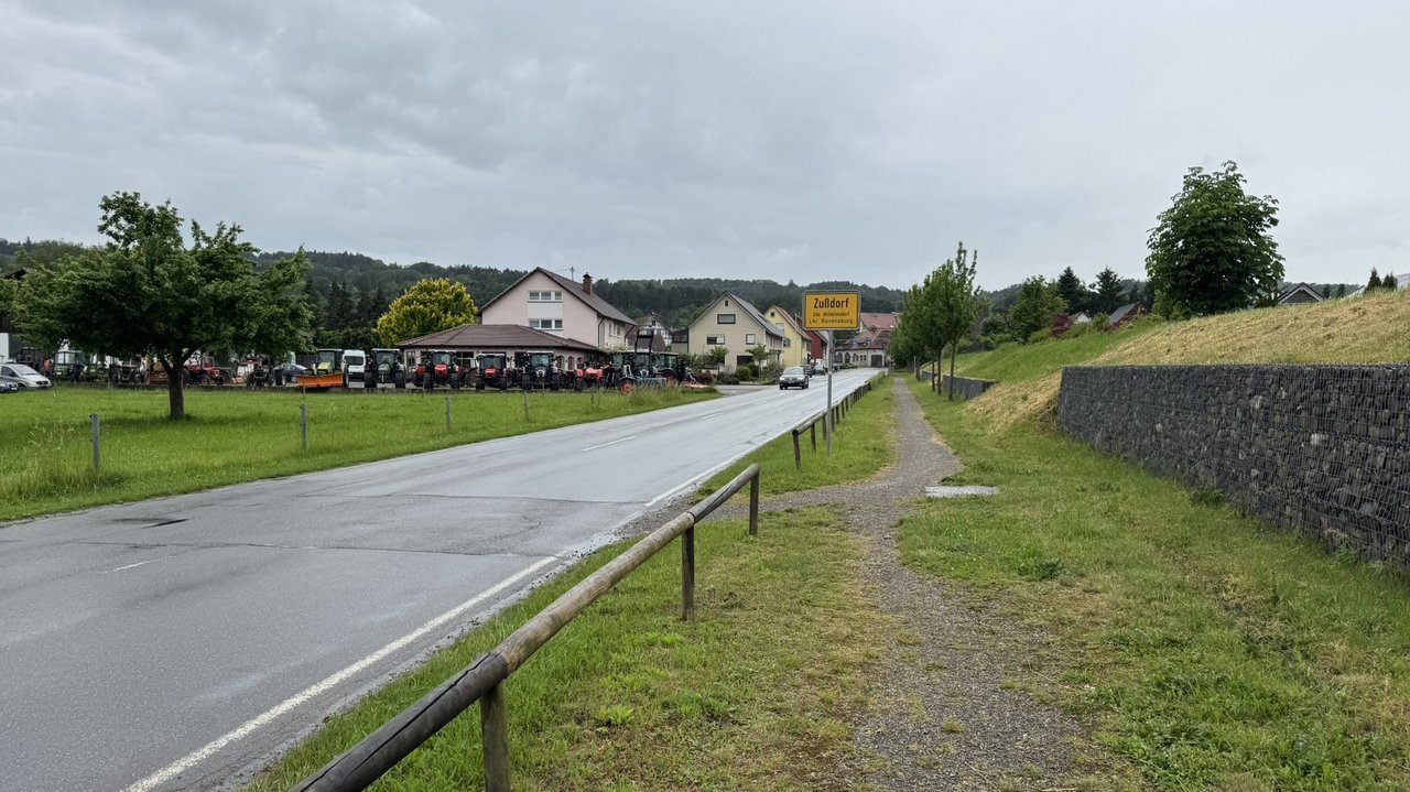 Das Foto zeigt die L 201b am Ortseingang Wilhelmsdorf-Zußdorf. Man sieht das Ortsschild und einige Gebäude. Auf der linken Seite stehen einige Traktoren.