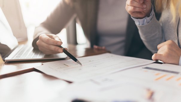 Zwei Kolleginnen arbeiten am Schreibtisch am Laptop und an Unterlagen, wie z.B. einer Statistik