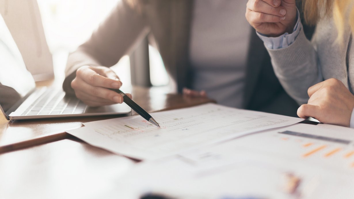 Zwei Kolleginnen arbeiten am Schreibtisch am Laptop und an Unterlagen, wie z.B. einer Statistik