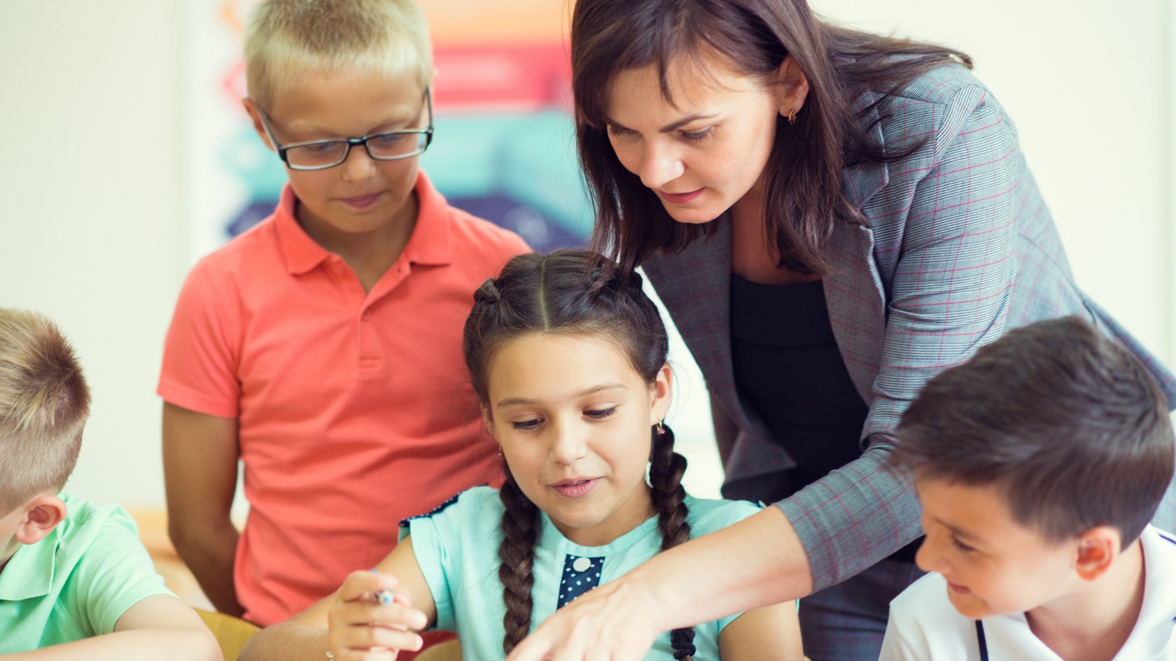 Ein Mädchen und vier Jungs und eine Lehrerin beugen sich über ein aufgeschlagenes Buch. Die Lehrerin zeigt auf den Text.