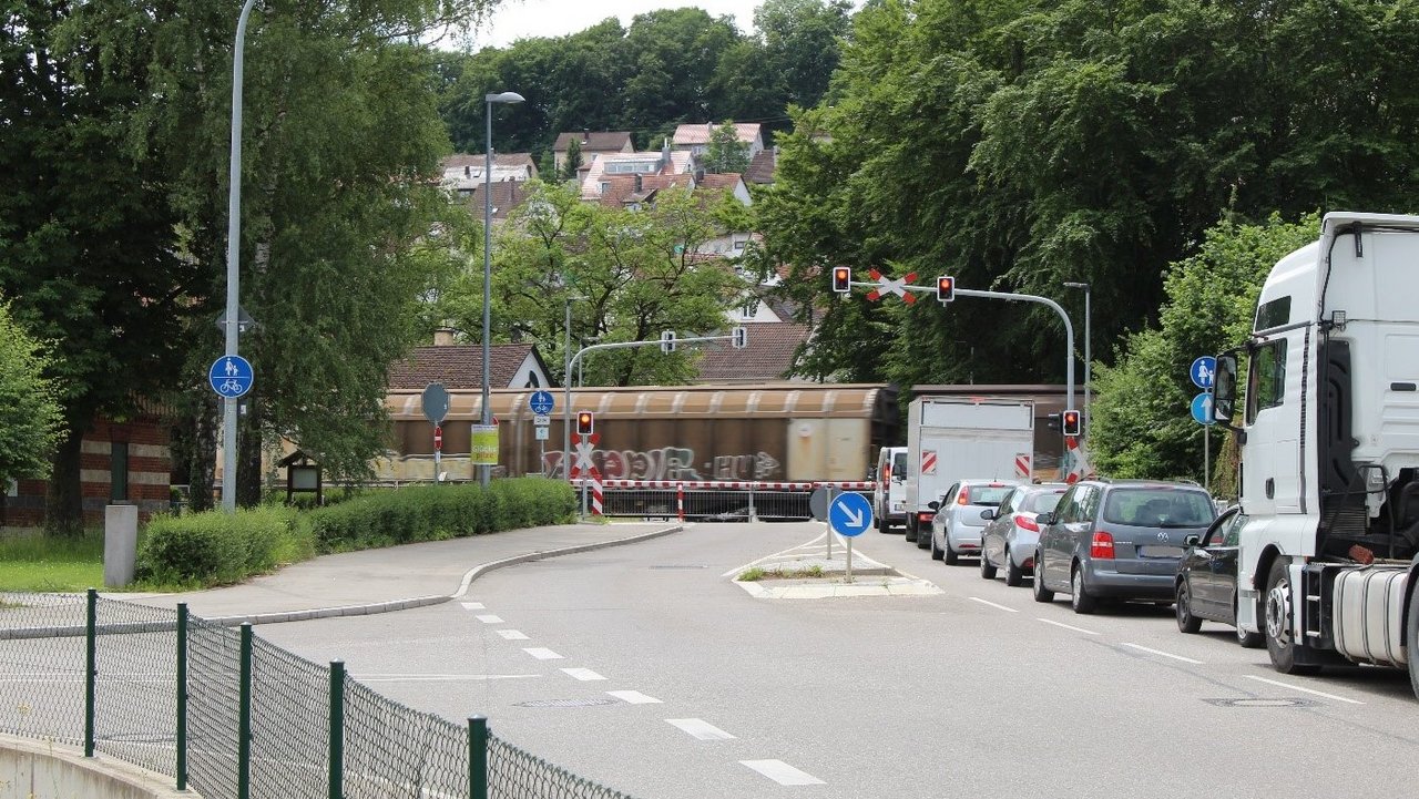 Das Foto zeigt einen Bahnübergang mit herunter gelassener Schranke. Mehrere Fahrzeuge stehen wartend davor. Im Hintergrund Wohngebäude von Blaustein-Klingelstein