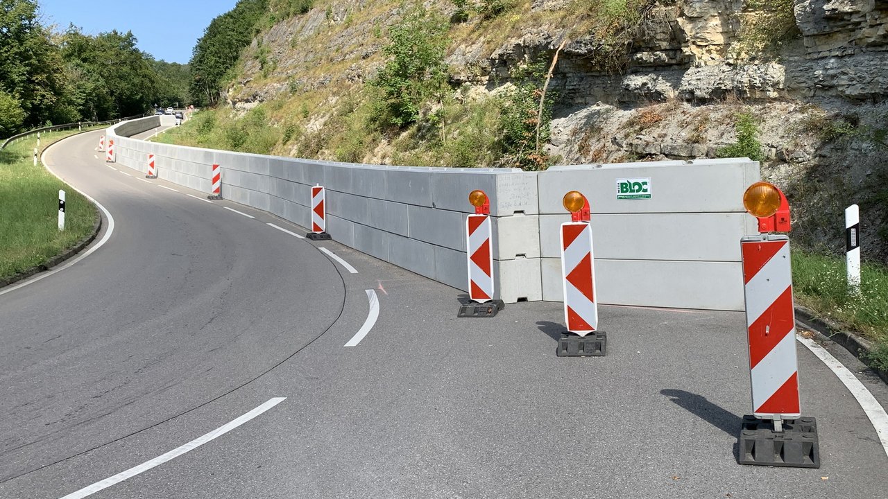 Das Foto zeigt die Hülbener Steige. Eine Wand aus Betonbauteilen sichert den rechten Streifen der Fahrbahn vor Felsstücken die vom Hang rutschen könnten. Mehrere Stehlen vor der Wand.