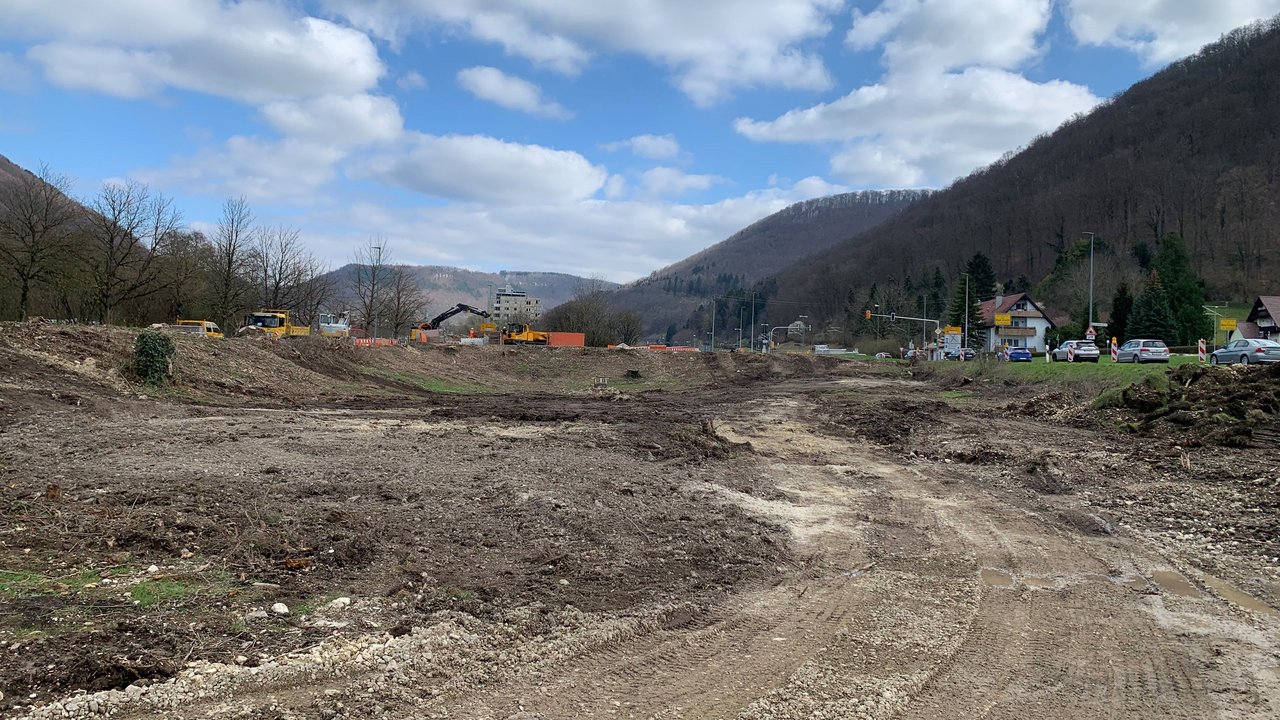Blick auf die künftige Baustelleneinrichtungsfläche mit mehreren Baustellenfahrzeugen. Rechts führt eine befahrene Straße vorbei.