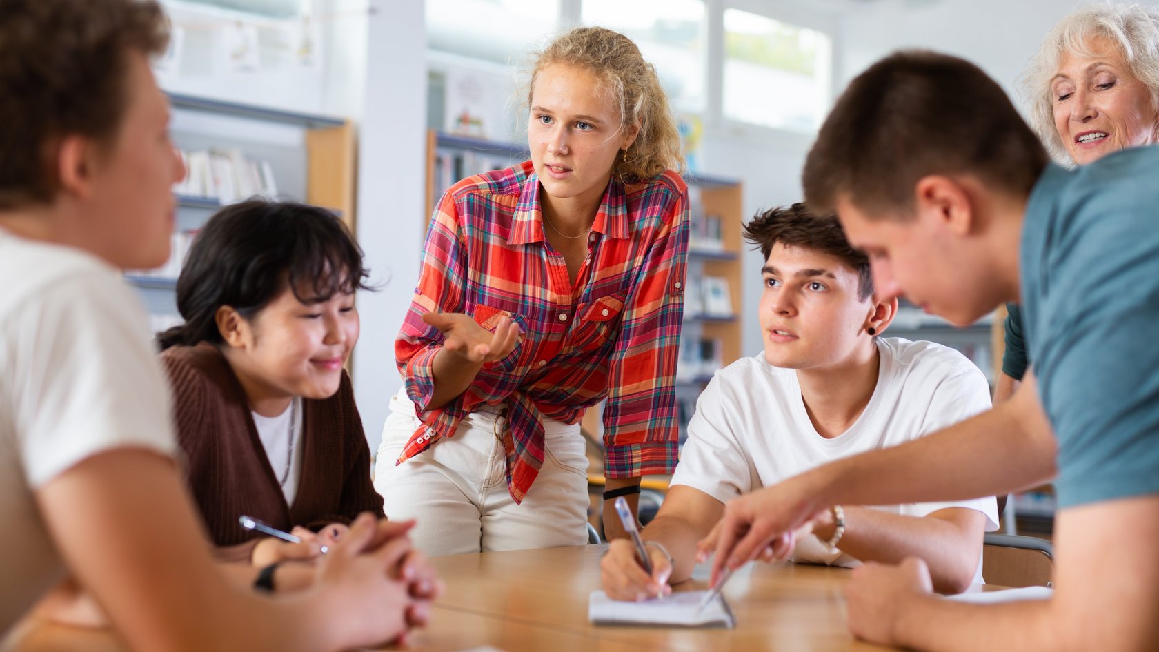 Schülerin und Schüler sitzen um einen runden Tisch bei der Gruppenarbeit unter Aufsicht einer Lehrerin