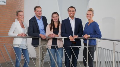 Drei junge Frauen und zwei Männer stehen an einem Geländer und lächeln in die Kamera