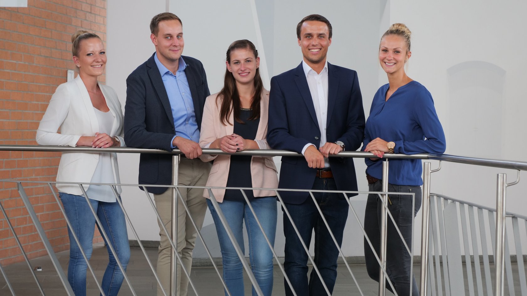 Drei junge Frauen und zwei Männer stehen an einem Geländer und lächeln in die Kamera