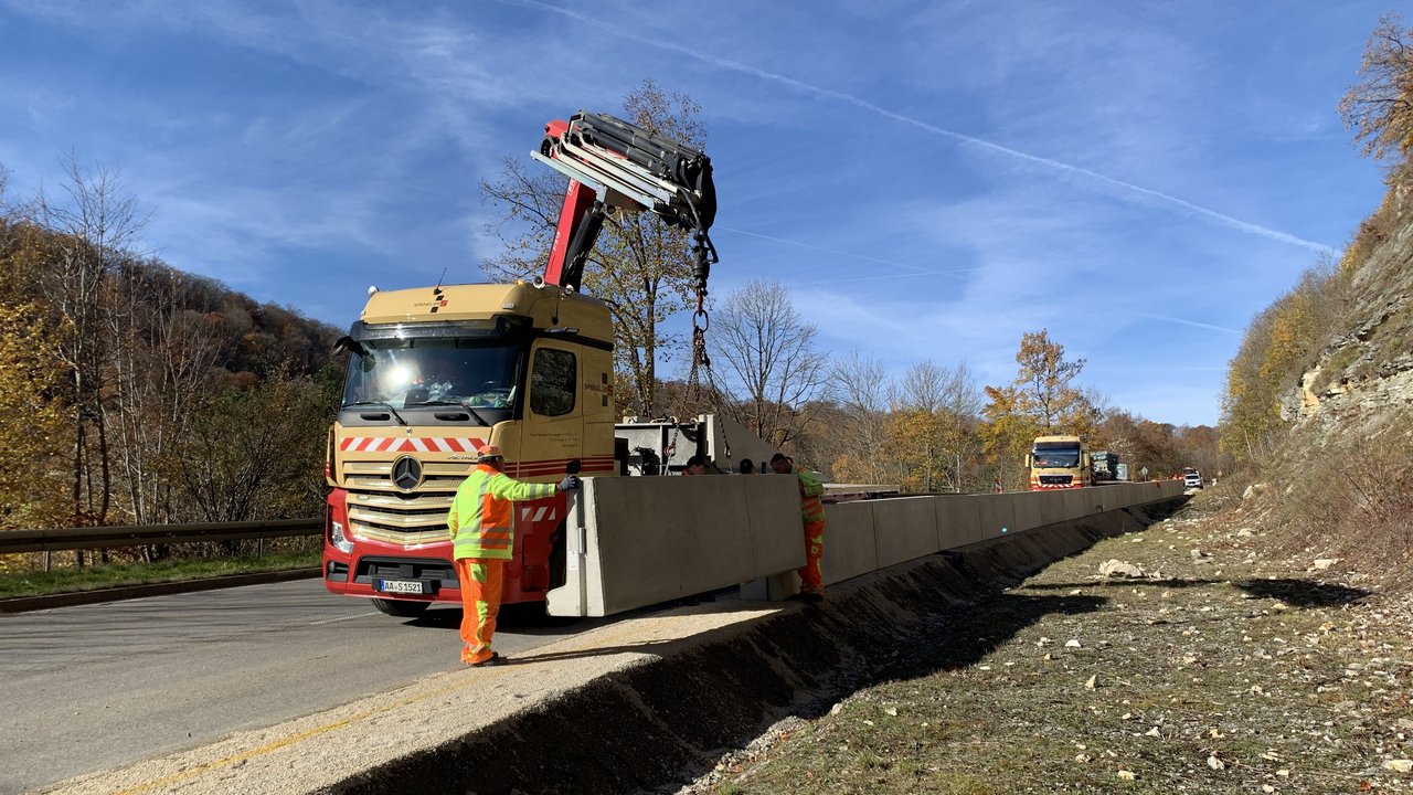 Das Foto zeigt einen Schwertransporter der mit einem Kran Betonfertigteilen zu einer Schwergewichtsmauer zur Felssicherung zusammenfügt.