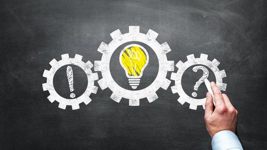 man sieht drei Zahnräder als Symbole für den Wirtschaftsmotor, im ersten sieht man ein Ausrufzeichen, im zweiten eine brennende Glühbirne, im dritten schreibt eine Hand ein Fragezeichen rein