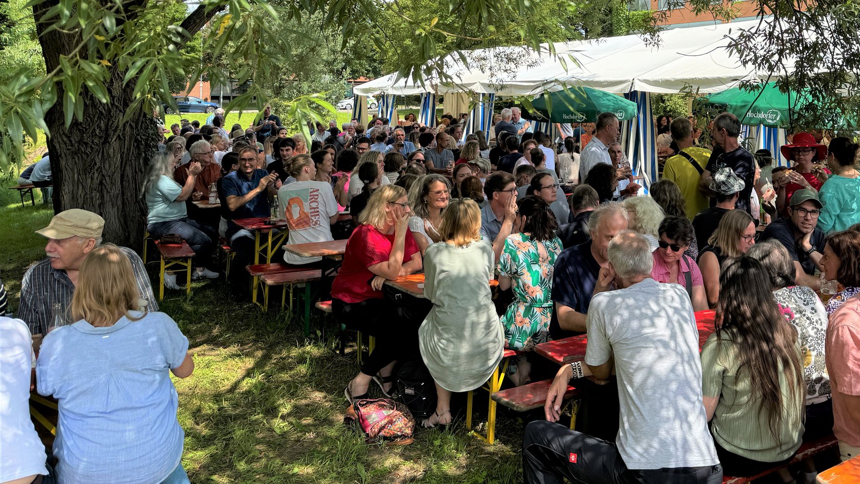 Menschen sitzen an Tischen und unterhalten sich. Im Hintergrund ist ein Festzelt zu sehen.