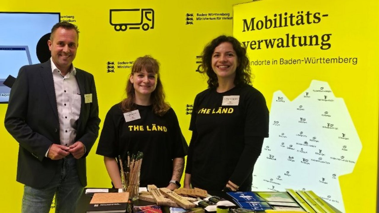 Das Foto zeigt einen Messestand mit gelben Stellwänden. 3 Personen stehen bei dem Messestand.