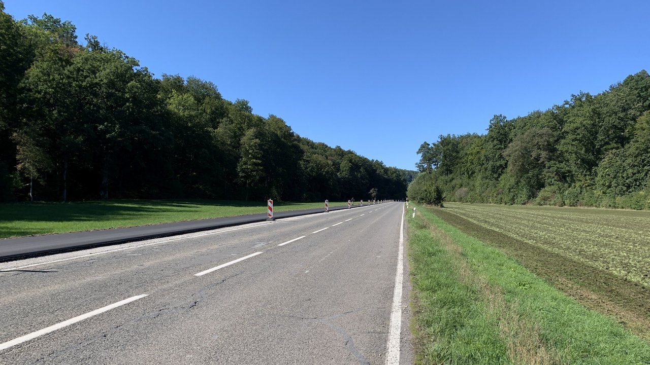 Man sieht die L 1208; links den neuen Radweg, Wiesen und Wald und rechts einen Acker und Wald bei blauem Himmel