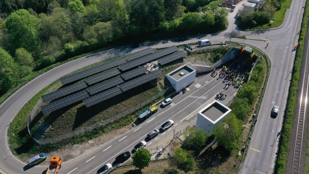 Drohnenfoto eines Straßen-Tunnelportals, vor dem viele Menschen auf der Straße stehen. Auf beiden Seiten der Straße sind kleine, fensterlose Neubauten. 