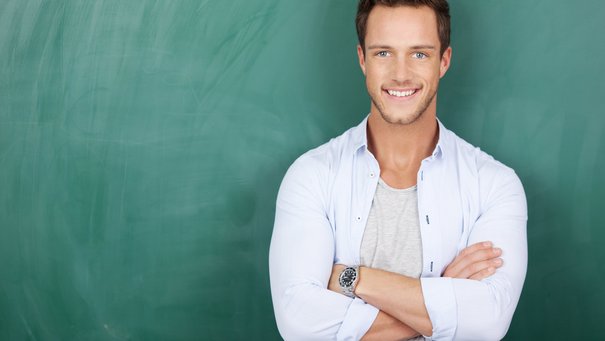 Junger Lehrer mit verschränkten Armen vor grüner Tafel