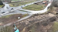 Ein Drohnenbild einer Kreuzung. Unten links sind ein Bagger und leere Gasleitungen zu sehen. An der Straße entlang sind viele Baumstümpfe. Im Hintergrund sind auf einer Wiese mehrere Steinhaufen zu sehen. 