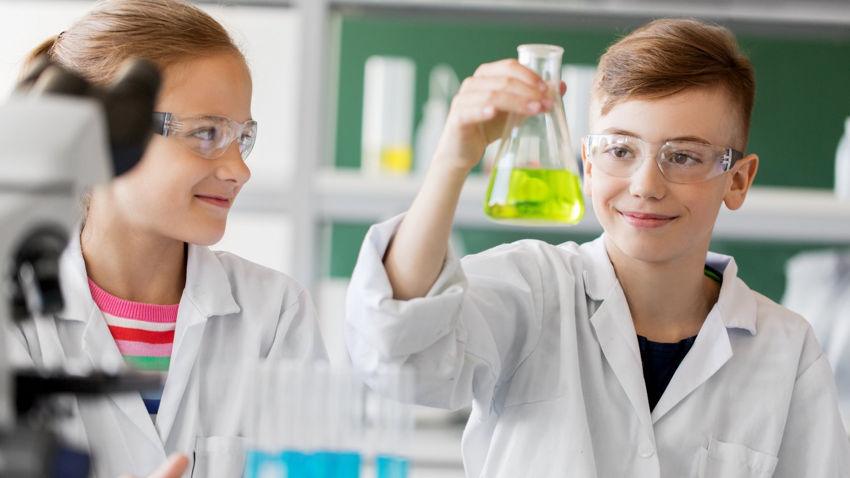 Zwei Kinder in Chemielabormänteln betrachten ein Reagenzglas mit grüner Flüssigkeit