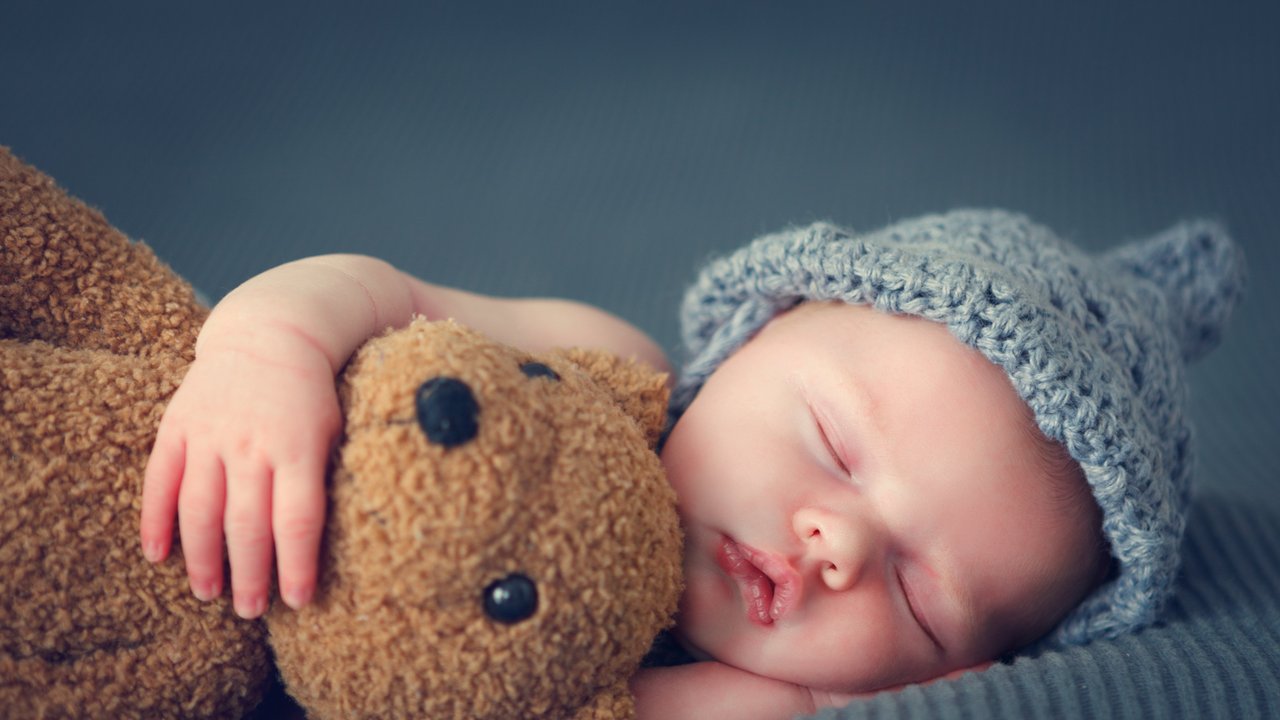 Man sieht ein schlafendes neugeborenes Baby mit einer blauen Mütze und einem Teddy im Arm