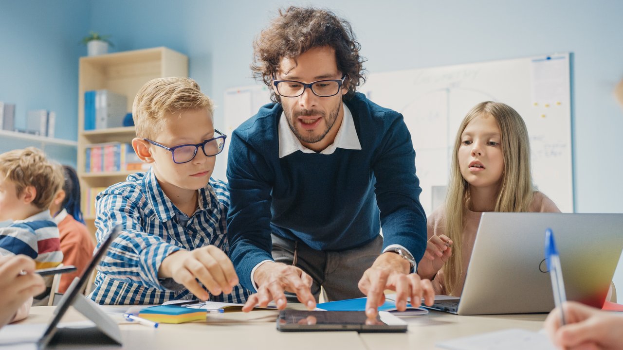 Ein Grundschüler und eine Grundschülerin sitzen am Tisch. Ein Lehrer beugt sich über sie und erklärt.