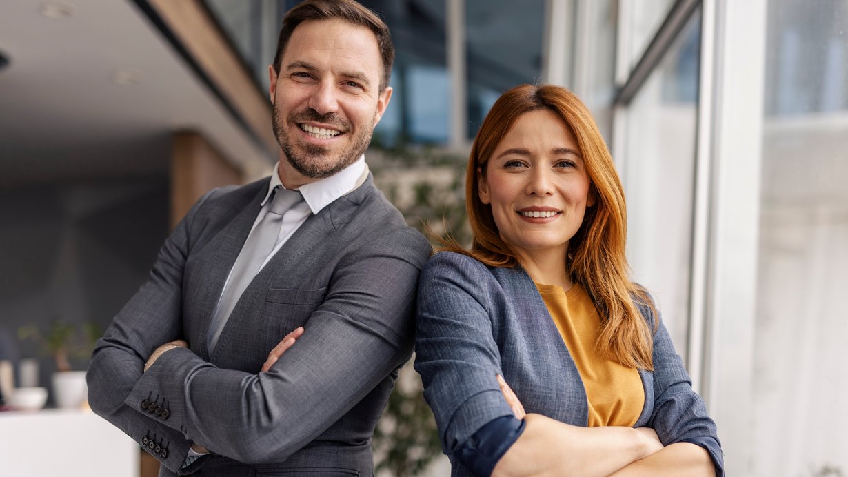 Ein Mann und eine Frau, beides Fachkräfte, lehnen mit verschränkten Armen aneinander und lächeln in die Kamera. 