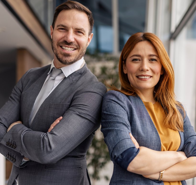 Ein Mann und eine Frau, beides Fachkräfte, lehnen mit verschränkten Armen aneinander und lächeln in die Kamera. 