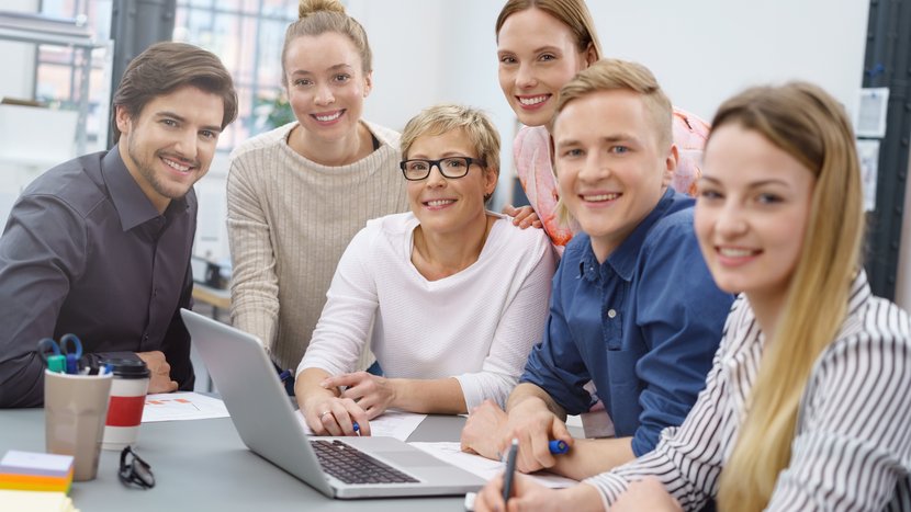 Eine gruppe aus Männern und Frauen unterschiedlichen Alters sitzen am Besprechungstisch und lächeln in die Kamera