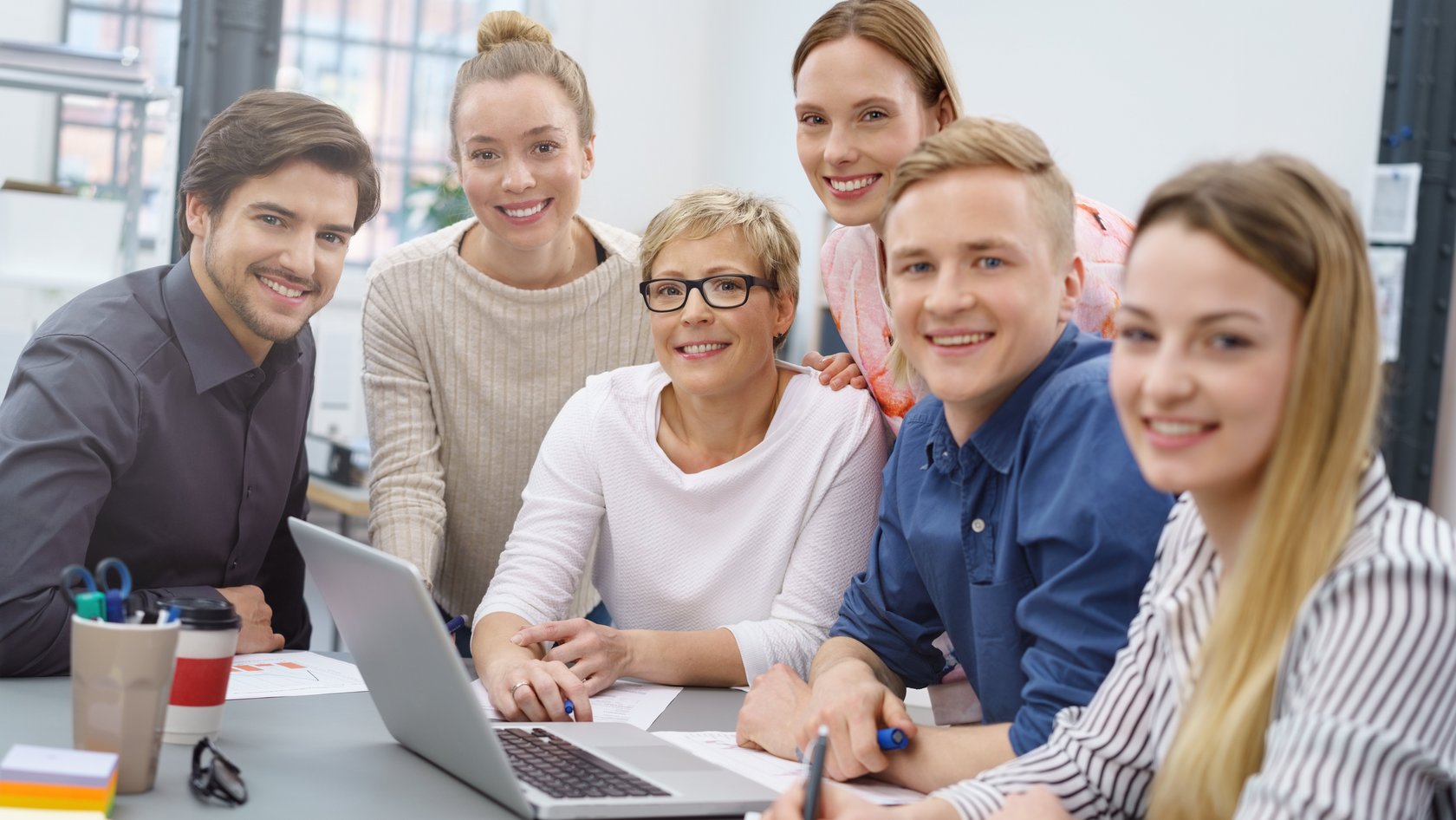 Eine gruppe aus Männern und Frauen unterschiedlichen Alters sitzen am Besprechungstisch und lächeln in die Kamera