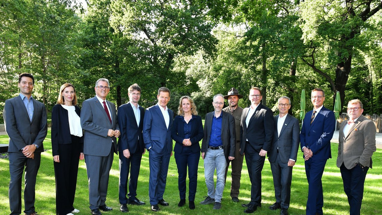 Das Bild zeigt 12 Personen bei einem Gruppenbild auf einer grünen Wiese; im Hintergrund Bäume