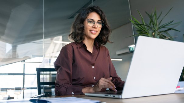 Eine junge Frau sitzt vor einem Laptop und lächelt in die Kamera.