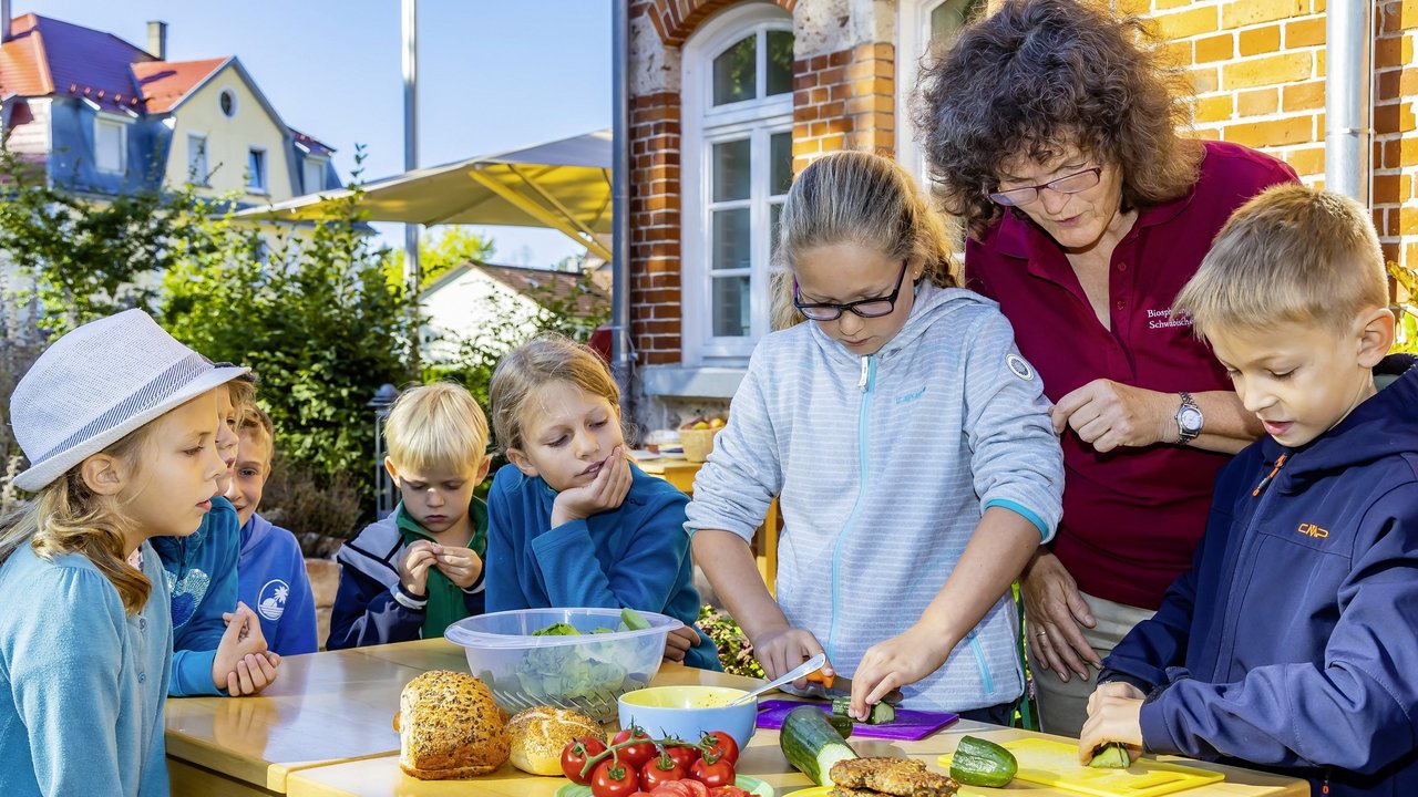 Das Foto zeigt mehrere Kinder und ein Frau, die an einem Tisch Gemüse schneiden