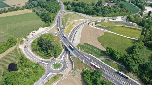 Luftbild einer Straße, die von links oben nach rechts unten verläuft. In der Mitte der Straße ist eine Brücke, unter der ein Kreisverkehr liegt. 