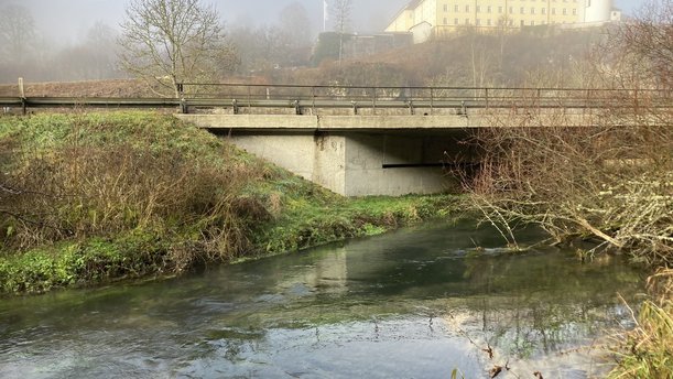 Eine Brücke, die einen Fluss überspannt, von der Seite. Im Hintergrund thront ein Kloster auf einem Hügel.
