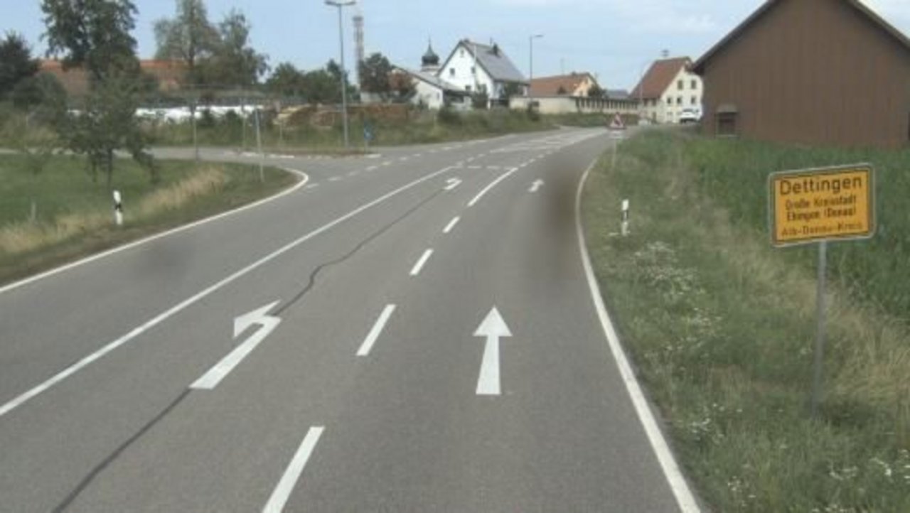 Das Bild zeigt eine Straße mit Fahrbahnmarkierungen und einer Abbiegespur nach links; Gebäude im Hintergrund