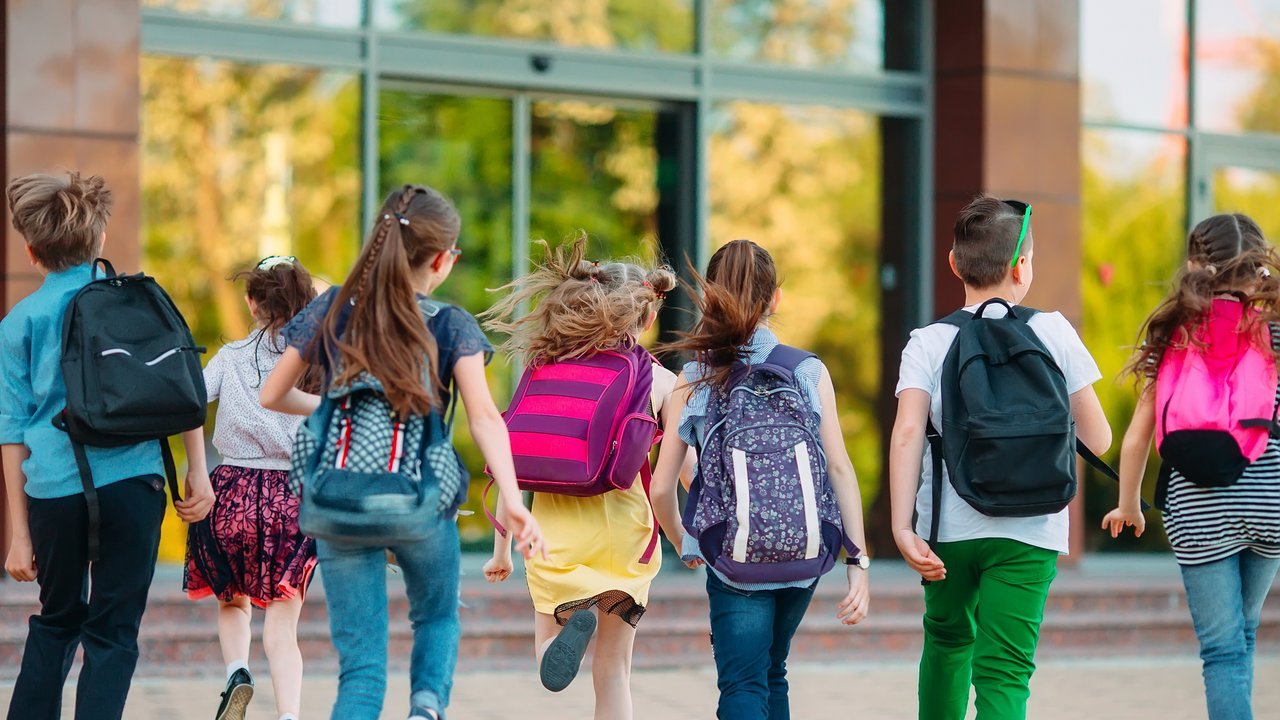7 Schulkinder mit Rucksäcken rennen auf ein Schulgebäude zu