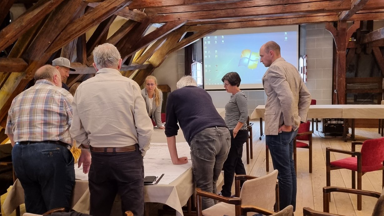 Fünf Männer und eine Frau stehen um einen Tisch, blicken auf Unterlagen und diskutieren miteinander. 