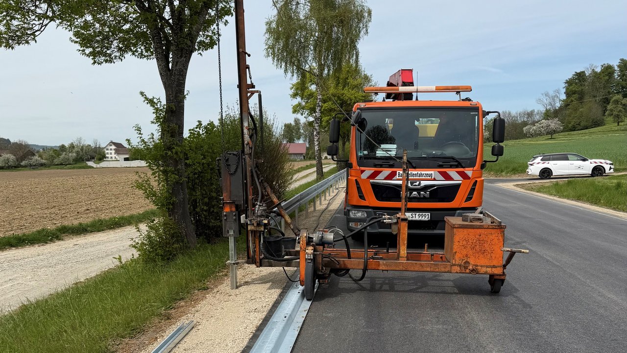 Das Foto zeigt ein Baustellenfahrzeug, das Schutzplankenarbeiten an einer Straße ausführt.
