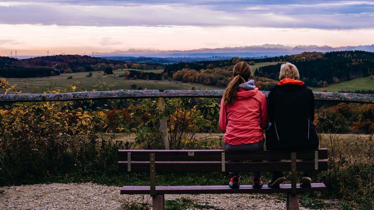 Das Bild zeigt zwei Personen von hinten sitzend auf einer Bank auf einem Berg, die den Ausblick auf eine Landschaft und den Sonnenaufgang betrachten.