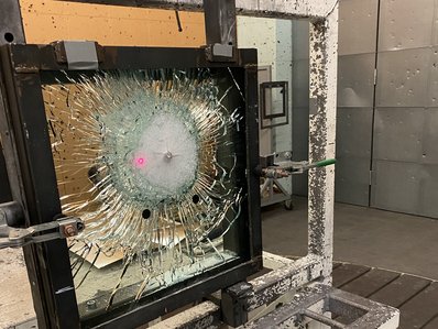 Eine Glasscheibe, die in einem Rahmen verschraubt ist, nach einem Beschuss. Die Scheibe ist zersplittert.