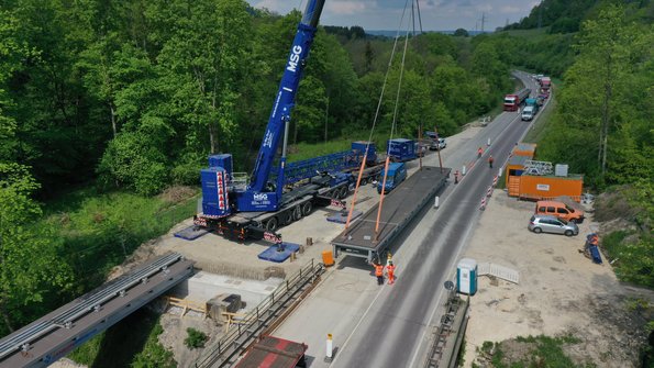Ein Kran steht neben einer Straße. Er Hebt ein Brückenteil, das direkt neben der Straße liegt, auf circa einen Meter Höhe. Vor dem Kran befinden sich Brücken-Widerlager, auf denen bereits ein Brückenteil abgelegt ist. 