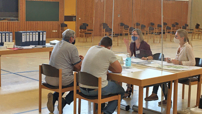eine Turnhalle mit Tischen, an einem Tisch sitzen 4 Personen im Gespräch