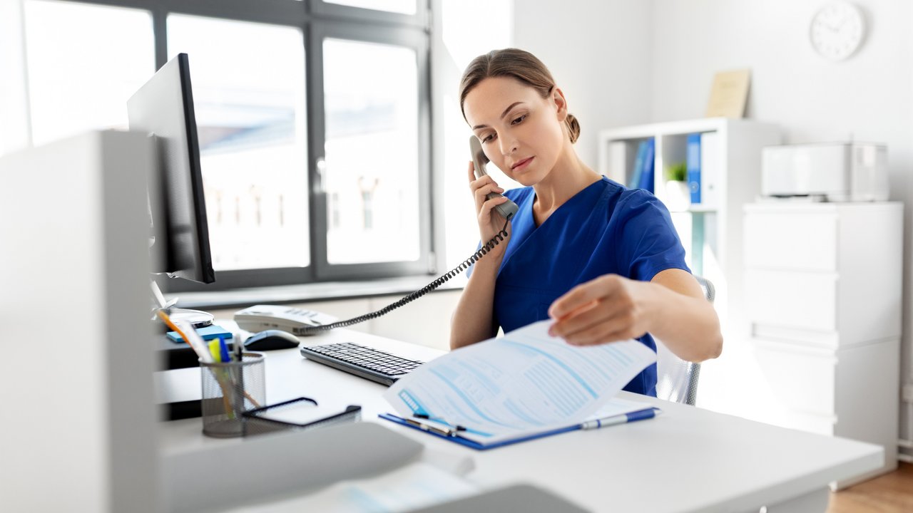 Eine Frau sitzt am Schreibtisch und blätter in Unterlagen während sie den Telefonhörer ans Ohr hält.