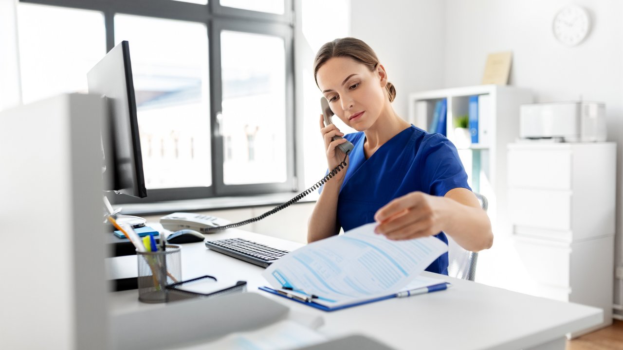 Eine Frau sitzt am Schreibtisch und blätter in Unterlagen während sie den Telefonhörer ans Ohr hält.