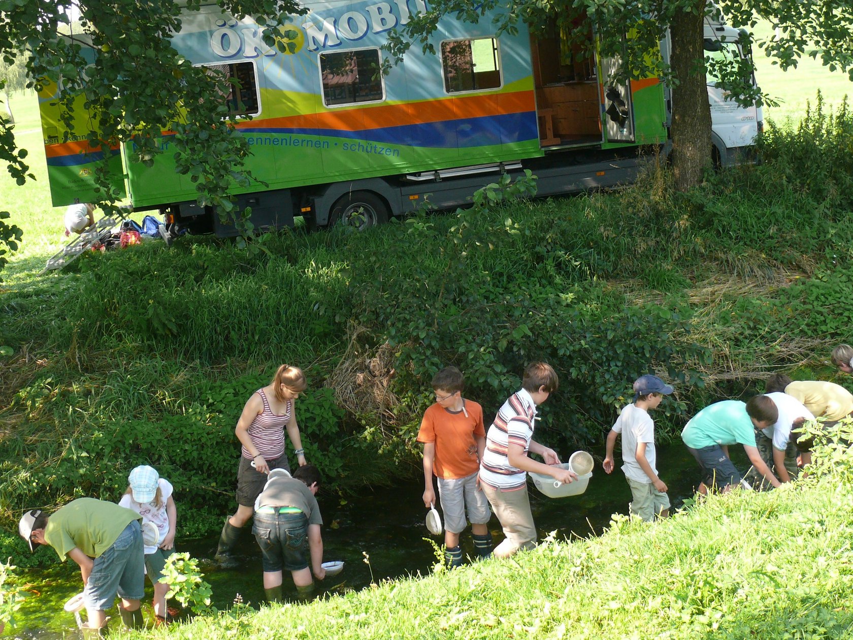 Kinder forschen im Bach - im Hintergrund sieht man das Ökomobil zwischen Bäumen