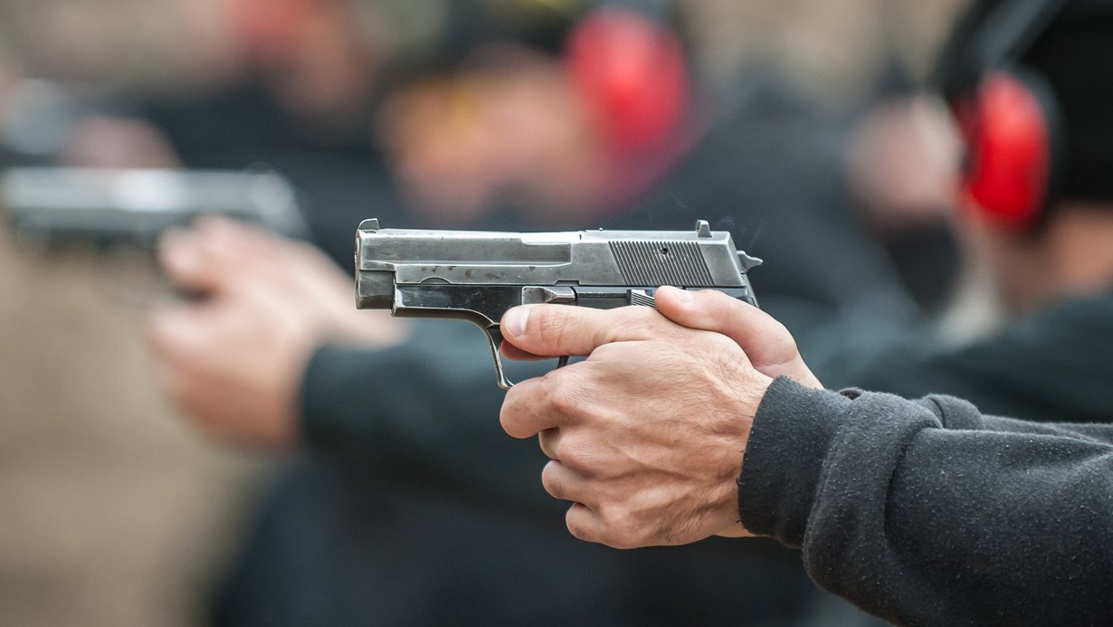 Sportschützen, die Schützen sieht man nur im Hintergrund, im Vordergrund ist eine Pistole, die abgefeuert wird