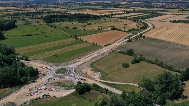 Luftbild einer Landschaft. Im unteren linken Viertel des Bildes ist ein Kreisverkehr zu sehen, der sich noch im Bau befindet: Der Kreisverkehr selbst ist asphaltiert, seine Anschlüsse noch nicht. Richtung rechts oben im Bild zieht sich vom Kreisverkehr ausgehend ein Erddamm. 