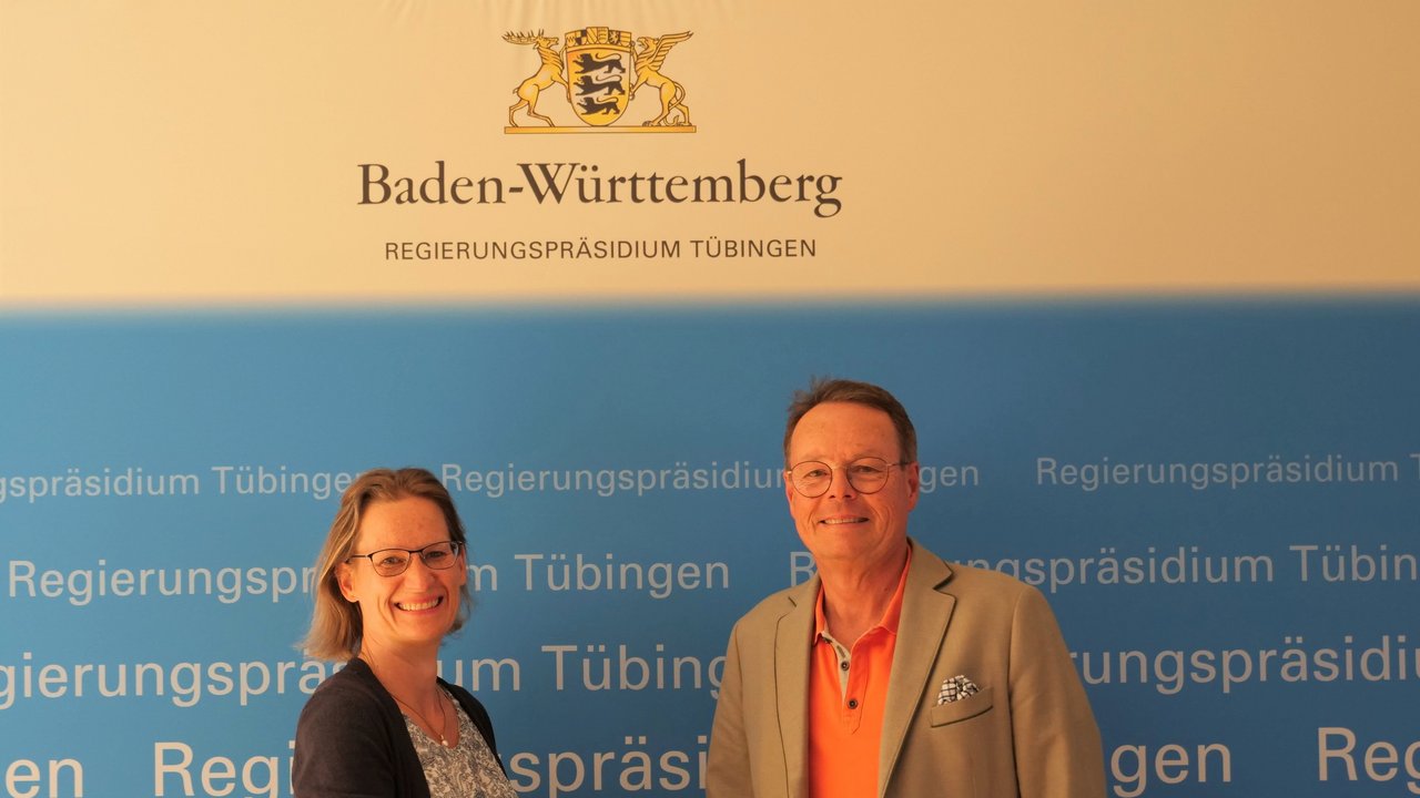 Das Foto zeigt Claudia Schneiderhan und Regierungspräsident Klaus Tappeser die sich die Hand reichen vor einer Plakatwand des RP Tübingen